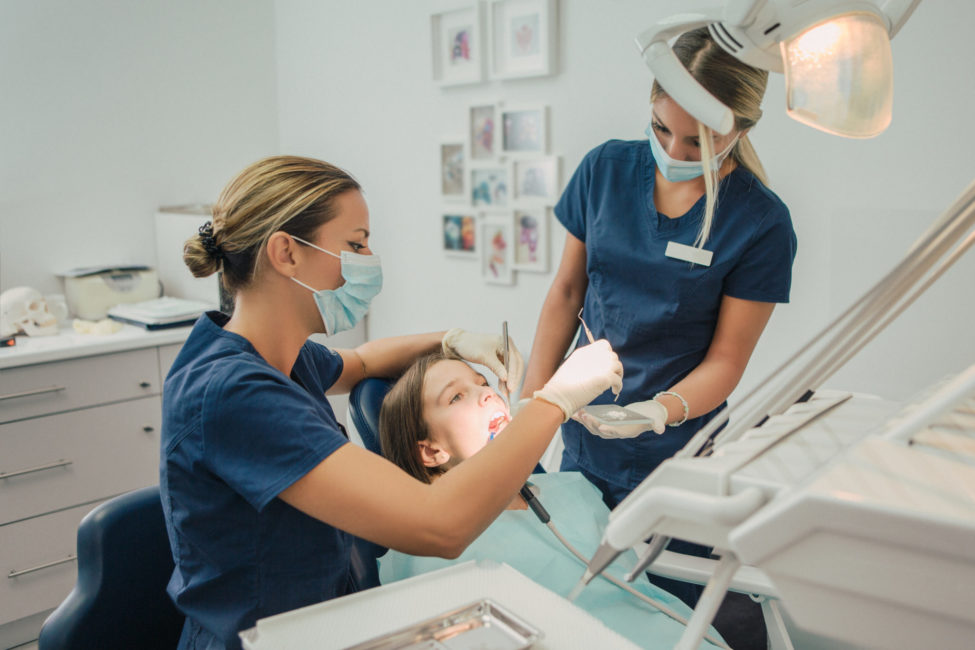 Kvinnelig tannhelsesekretær som får hjelp av en kvinnelig kollega til å undersøke tennene til en pasient som sitter i tannlegestolen.